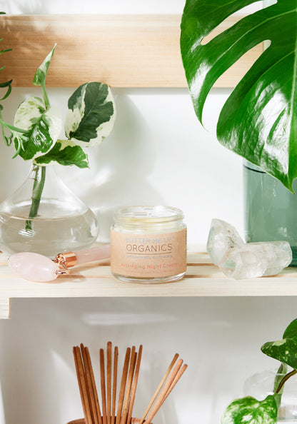 A jar of "White Smokey Anti Aging Night Cream Face Moisturizer Organic" on a shelf surrounded by green plants, crystals, and incense sticks, conveying a serene spa-like atmosphere. This anti-wrinkle cream is formulated with all-natural ingredients.