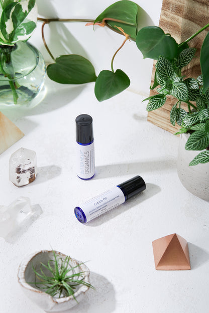 Two bottles of White Smokey Organic Cuticle Oil on a white surface surrounded by plants, crystals, and geometric decor elements. Natural light illuminates the scene.