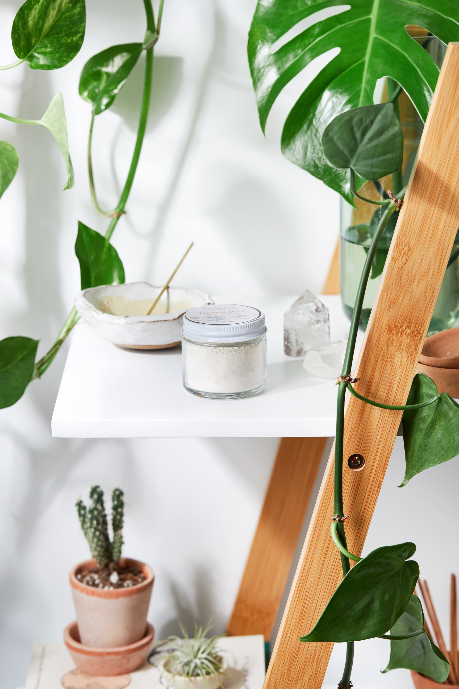 A jar of White Smokey Turmeric Mask cosmetic product on a white shelf with natural decor elements like plants and a piece of crystal.