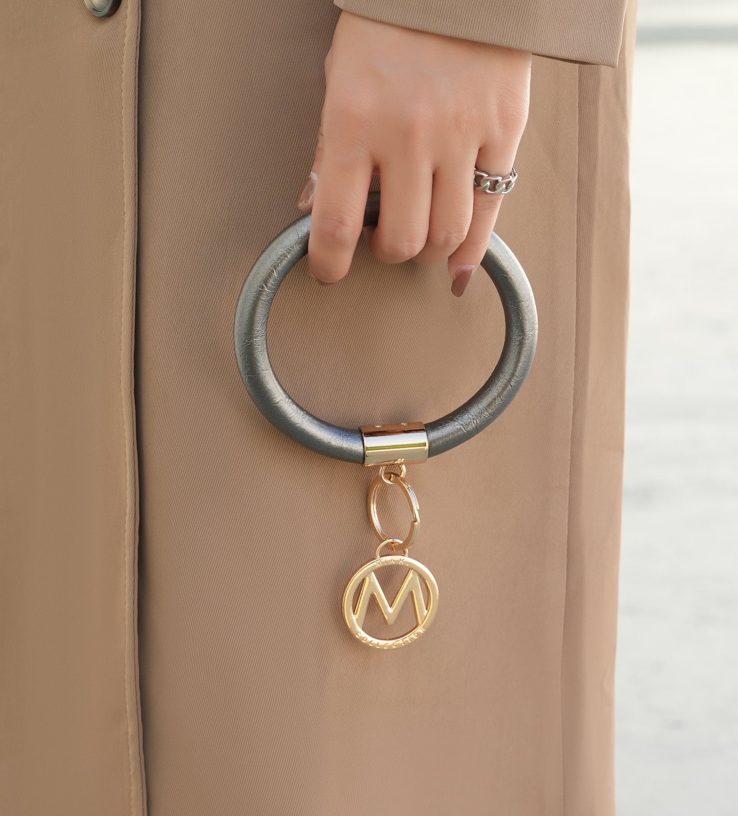 Close-up of a person's hand gripping a large Jasmine Vegan Leather Women Bangle Wristlet Keychain set attached to a beige Pink Orpheus coat with a golden 'm' pendant.