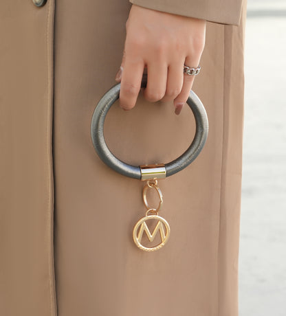 Close-up of a person's hand gripping a large Jasmine Vegan Leather Women Bangle Wristlet Keychain set attached to a beige Pink Orpheus coat with a golden 'm' pendant.