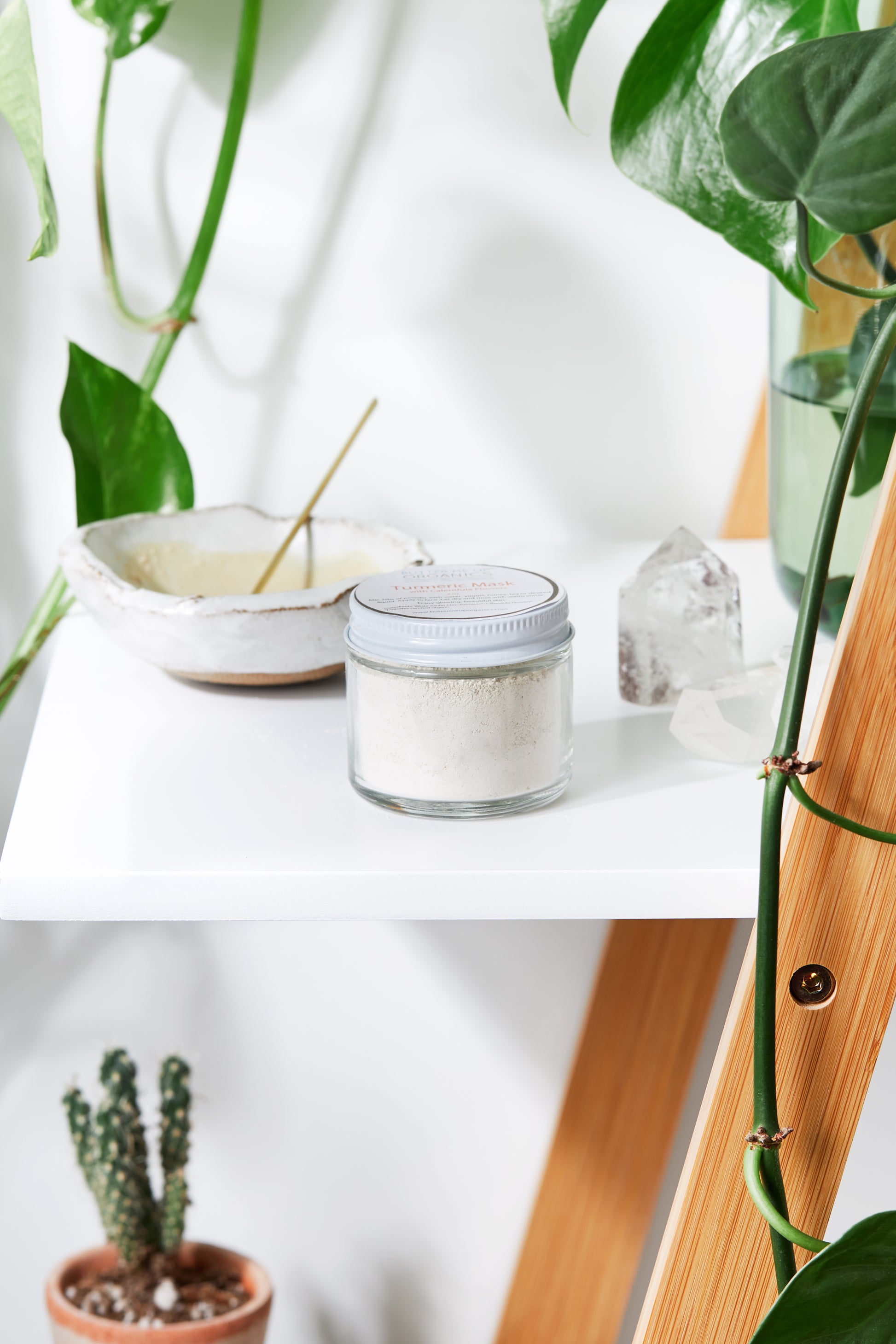 Glass jar of White Smokey Turmeric Mask on a wooden shelf surrounded by green plants and natural decor elements.