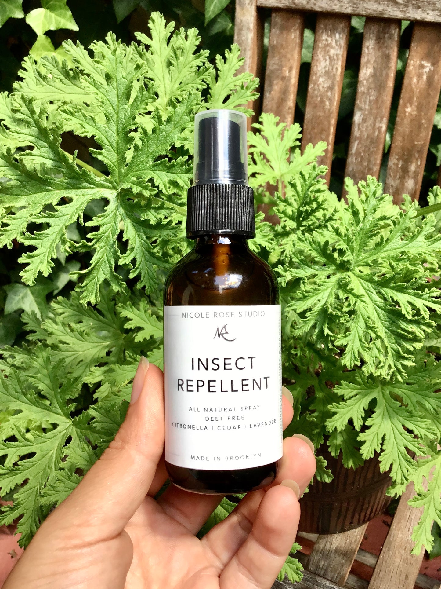 A hand holding a bottle of Indigo Poseidon's All Natural Insect Repellent, crafted with essential oils and DEET-free, is displayed in front of a green plant on a wooden chair.