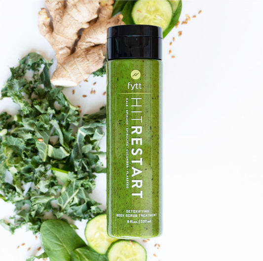 A bottle of Cyan Ares hit restart detoxifying exfoliating mask surrounded by slices of cucumber, ginger, and kale leaves on a white background.