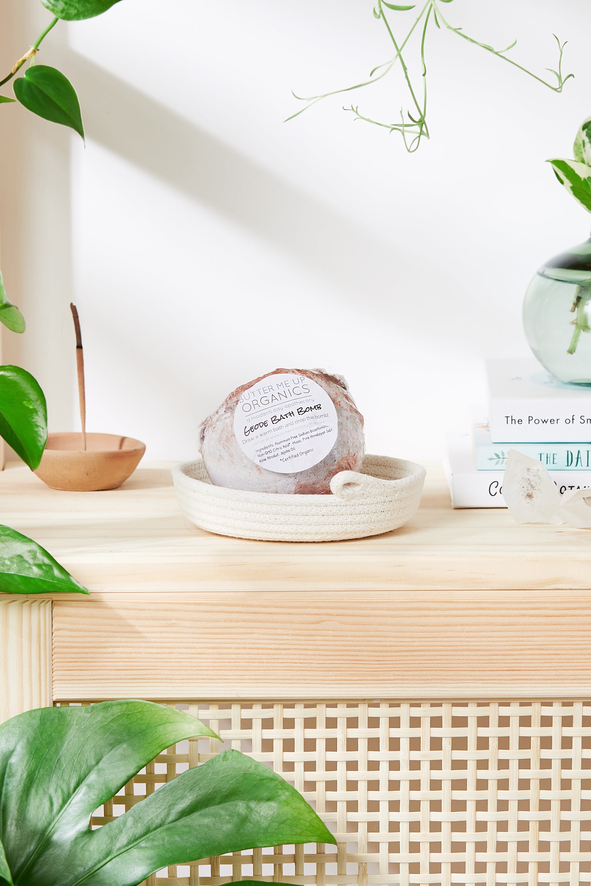 A soap bar labeled "Organic Geode Bath Bombs" from White Smokey rests on a woven dish atop a light wooden surface. Surrounding items include books, a small plant, and an incense holder with Himalayan salt, set against a white background with greenery.