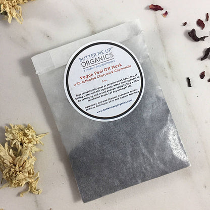 A packet labeled "White Smokey VEGAN activated charcoal peel off mask" is placed on a marble surface next to scattered dried flowers, highlighting its vegan version as an effective blackhead removal solution.