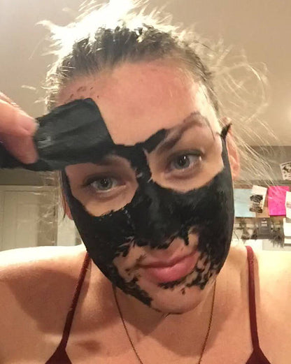 A person wearing a White Smokey VEGAN activated charcoal peel off mask is peeling it off from their forehead using their hand. The background shows a room with various items and notes on the wall, suggesting an environment where skincare routines are essential.