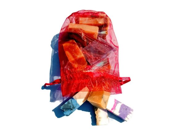 A red drawstring mesh bag containing a variety of colorful, rectangular handmade soap bars from the Yellow Shadow brand's Assorted Vegan Soap Sampler - 1lb, displayed on a white background. Enjoy an unparalleled bath experience with this exquisite vegan soap collection, each piece crafted to delight your senses.