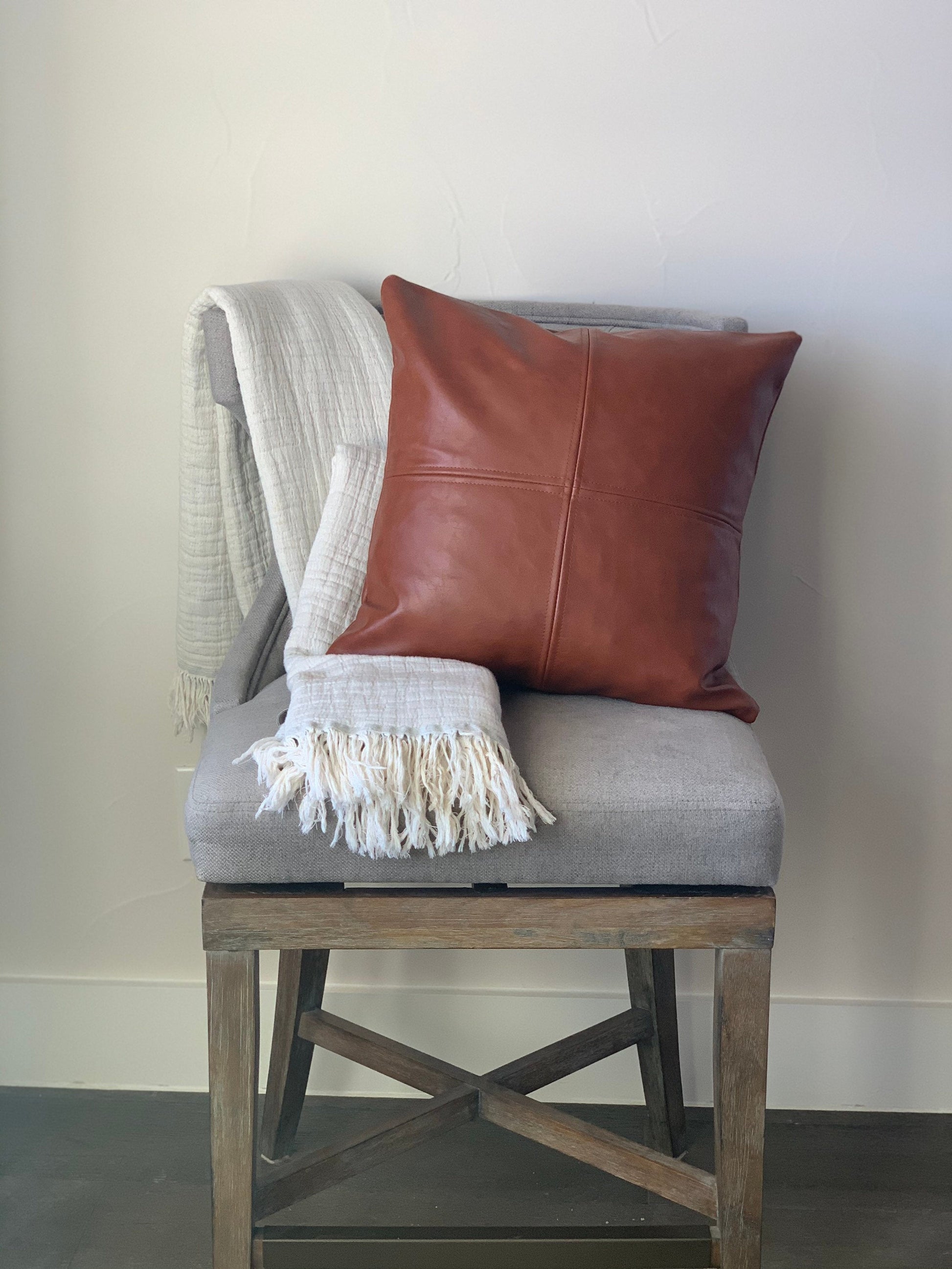 A gray chair with a large brown Magenta Charlie vegan leather pillow cover and a draped white throw blanket with fringe detailing.