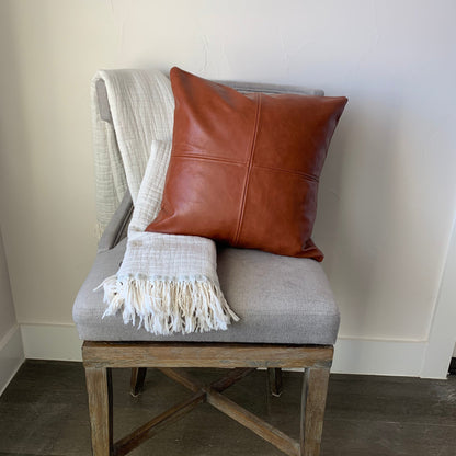A brown Magenta Charlie vegan leather pillow cover on a gray chair, partially covered by a light beige Magenta Charlie throw with fringe, against a white wall.