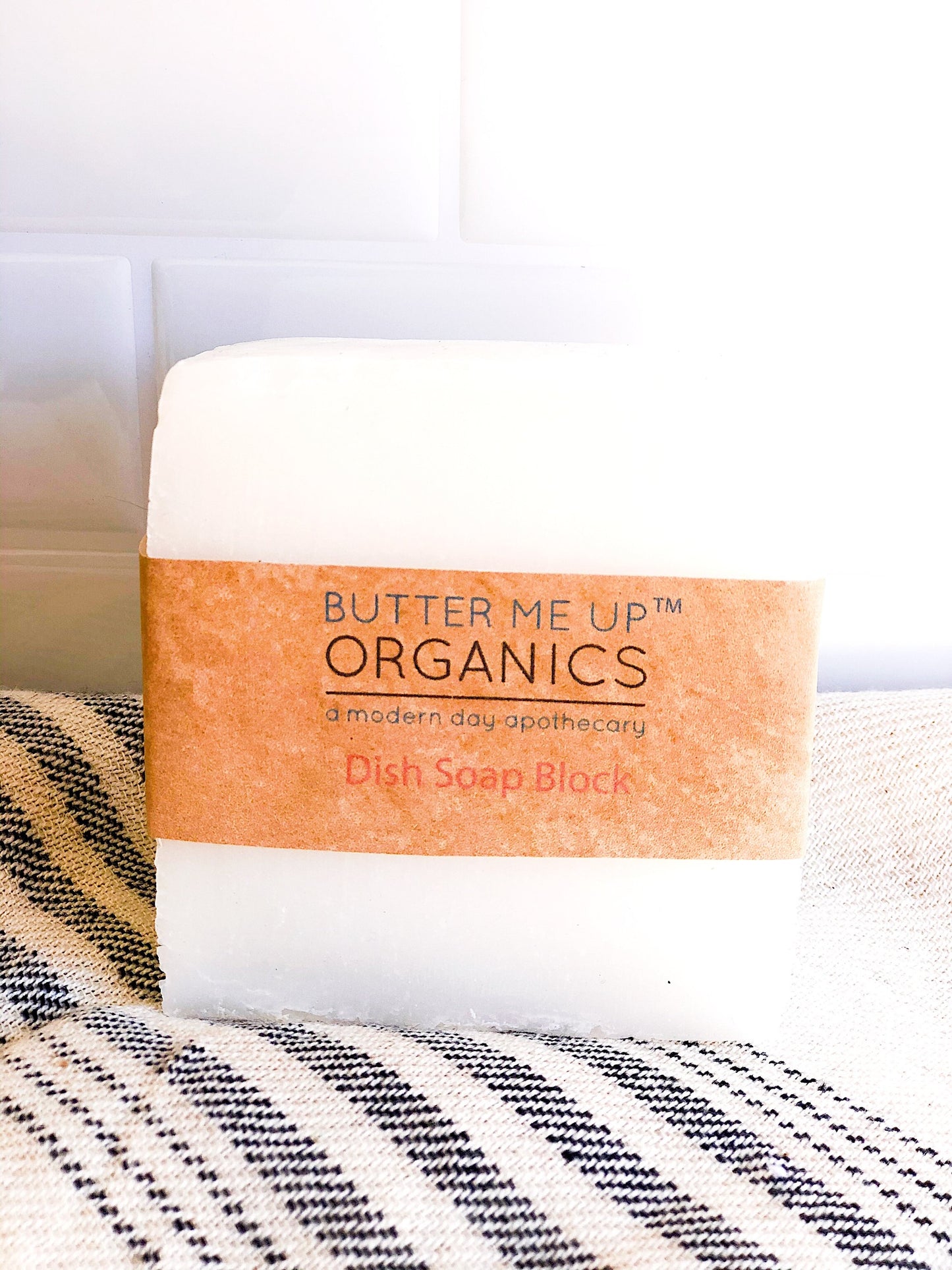 A white block of Organic Solid Dish Soap from White Smokey, featuring a brown paper label, rests on a striped cloth. This eco-friendly product promotes zero waste living while adding charm to your kitchen essentials.