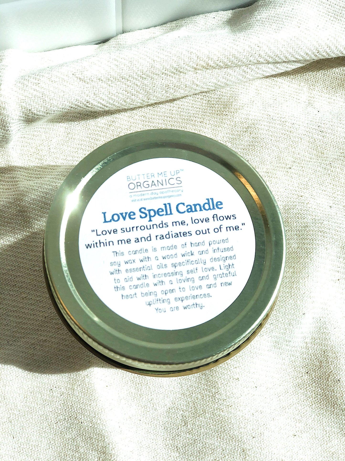 A small, round "White Smokey" Love Spell intention candle in a tin, displayed in sunlight, with an inspirational quote printed on its label.