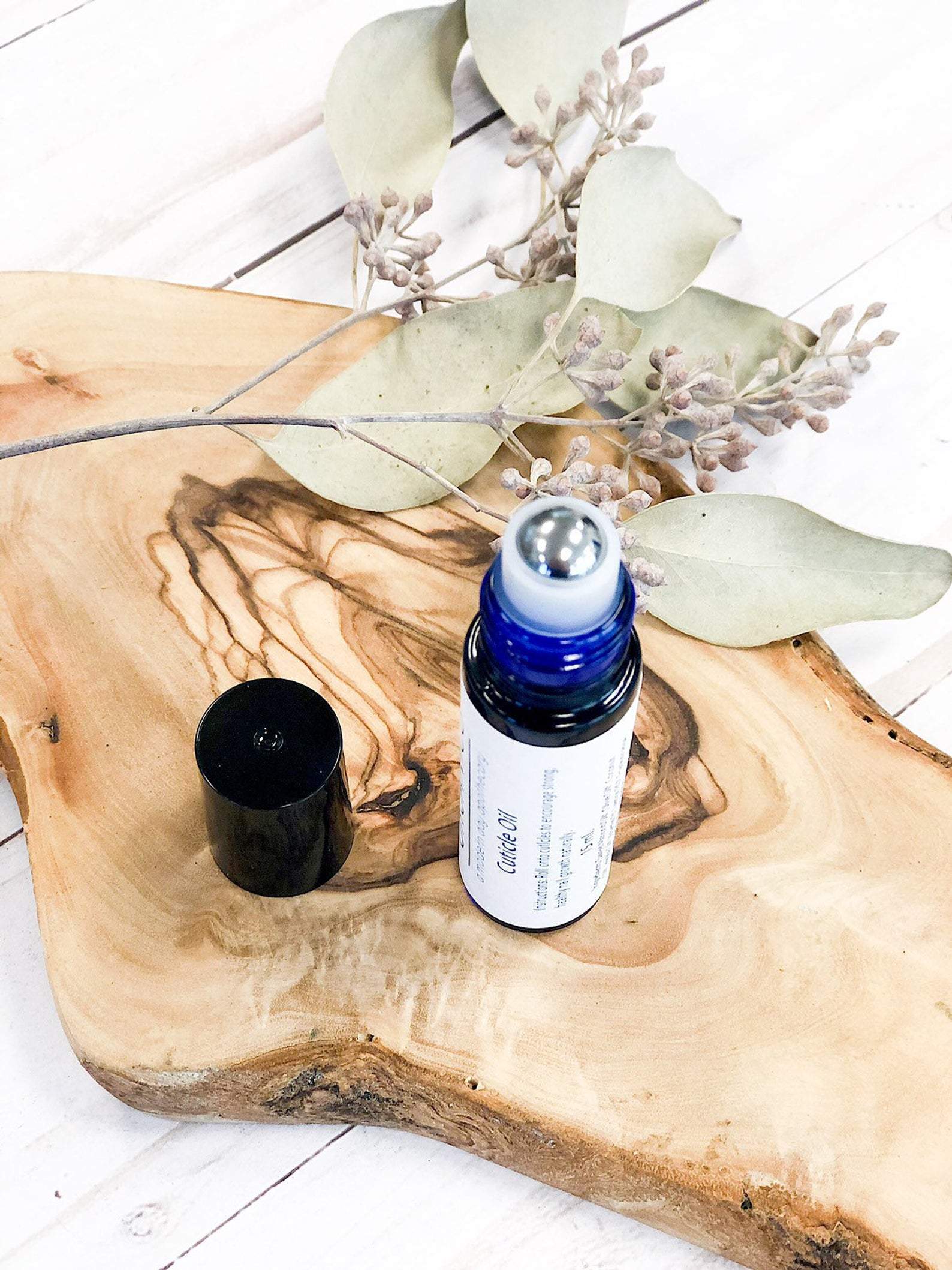 A blue glass roll-on bottle of White Smokey Organic Cuticle Oil, featuring a black cap next to a eucalyptus branch on a wooden slab with a swirling grain pattern.