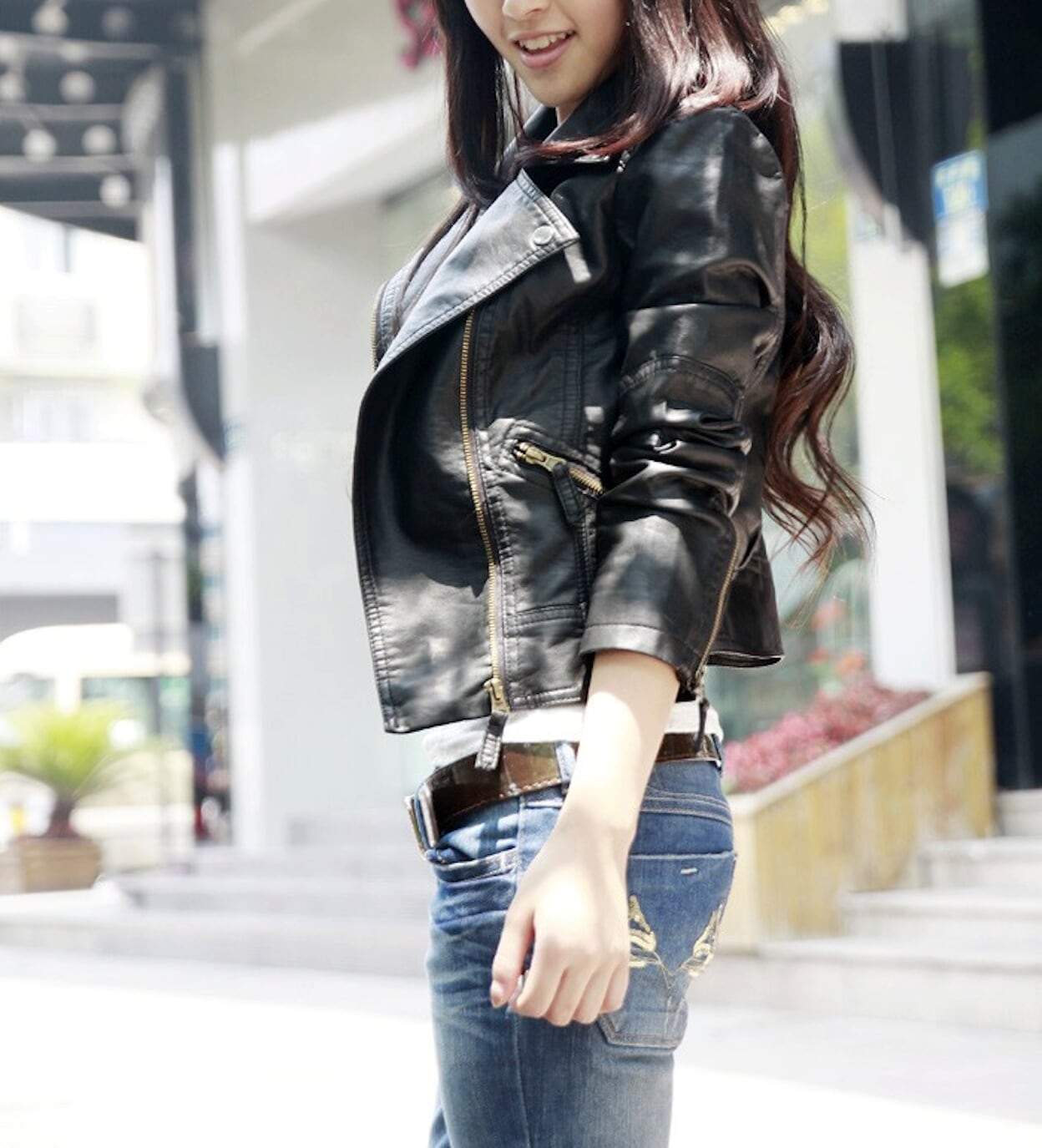 A person wearing a black Womens Cropped Vegan Leather Jacket by Yellow Pandora and blue jeans stands outdoors near a building.