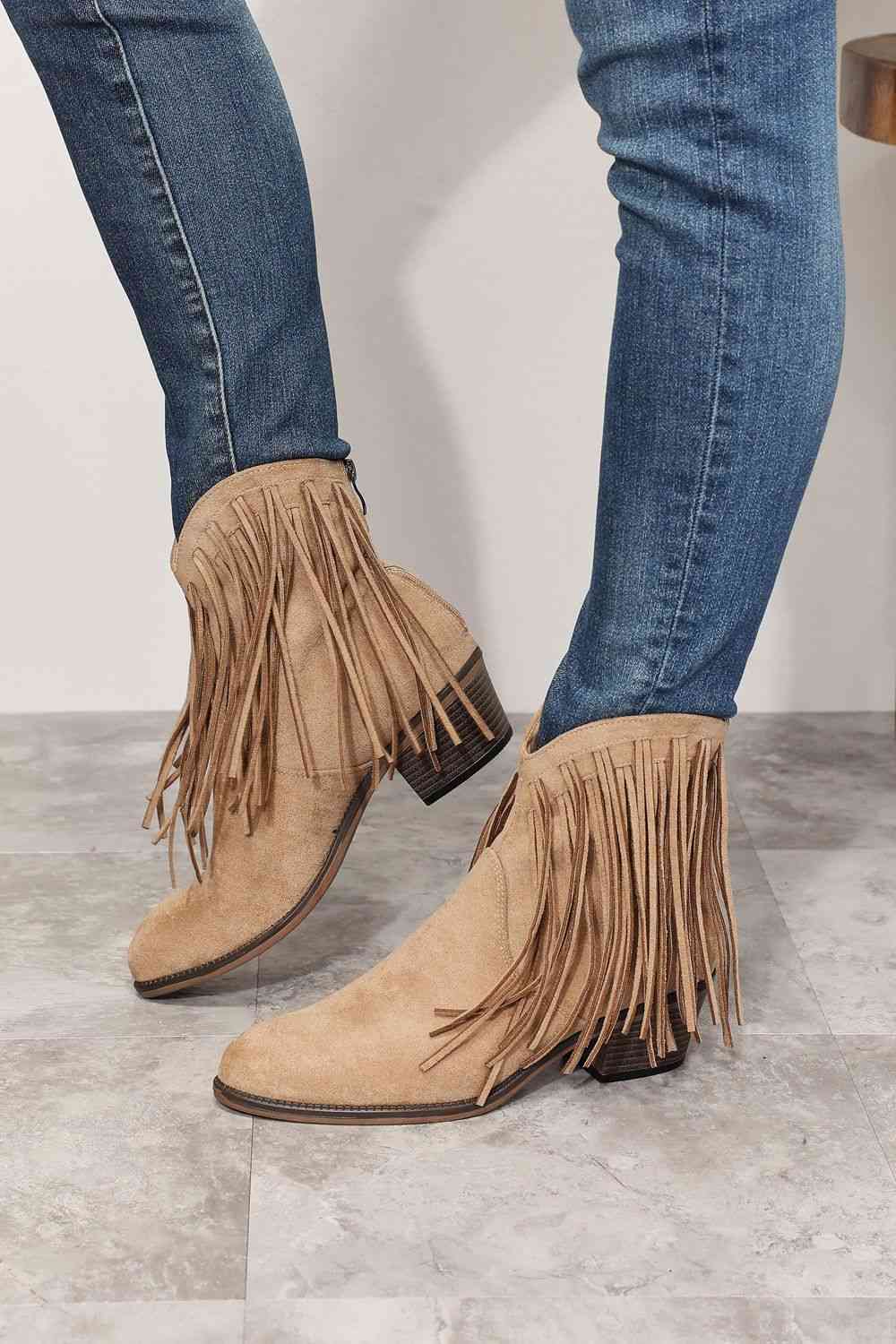 A person wearing Trendsi Legend Women's Fringe Cowboy Western Ankle Boots with long fringe details, paired with blue jeans, standing on a tiled floor.