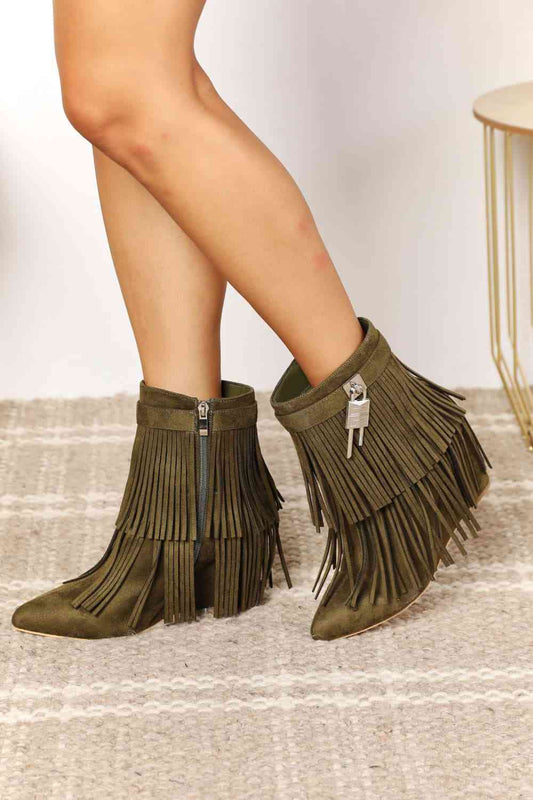 A pair of Legend Women's Tassel Wedge Heel Ankle Booties in olive green on a woman's feet, displayed against a neutral-toned background with a textured rug.