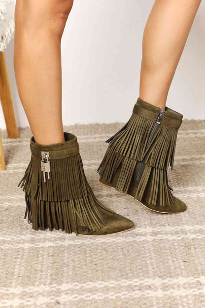 A person wearing green vegan suede Legend Women's Tassel Wedge Heel ankle boots with fringe detail, standing on a woven rug from Trendsi.