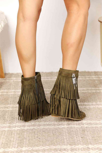 A person wearing green vegan suede Legend Women's Tassel Wedge Heel Ankle Booties with long fringe detail, standing on a textured rug against a beige backdrop by Trendsi.