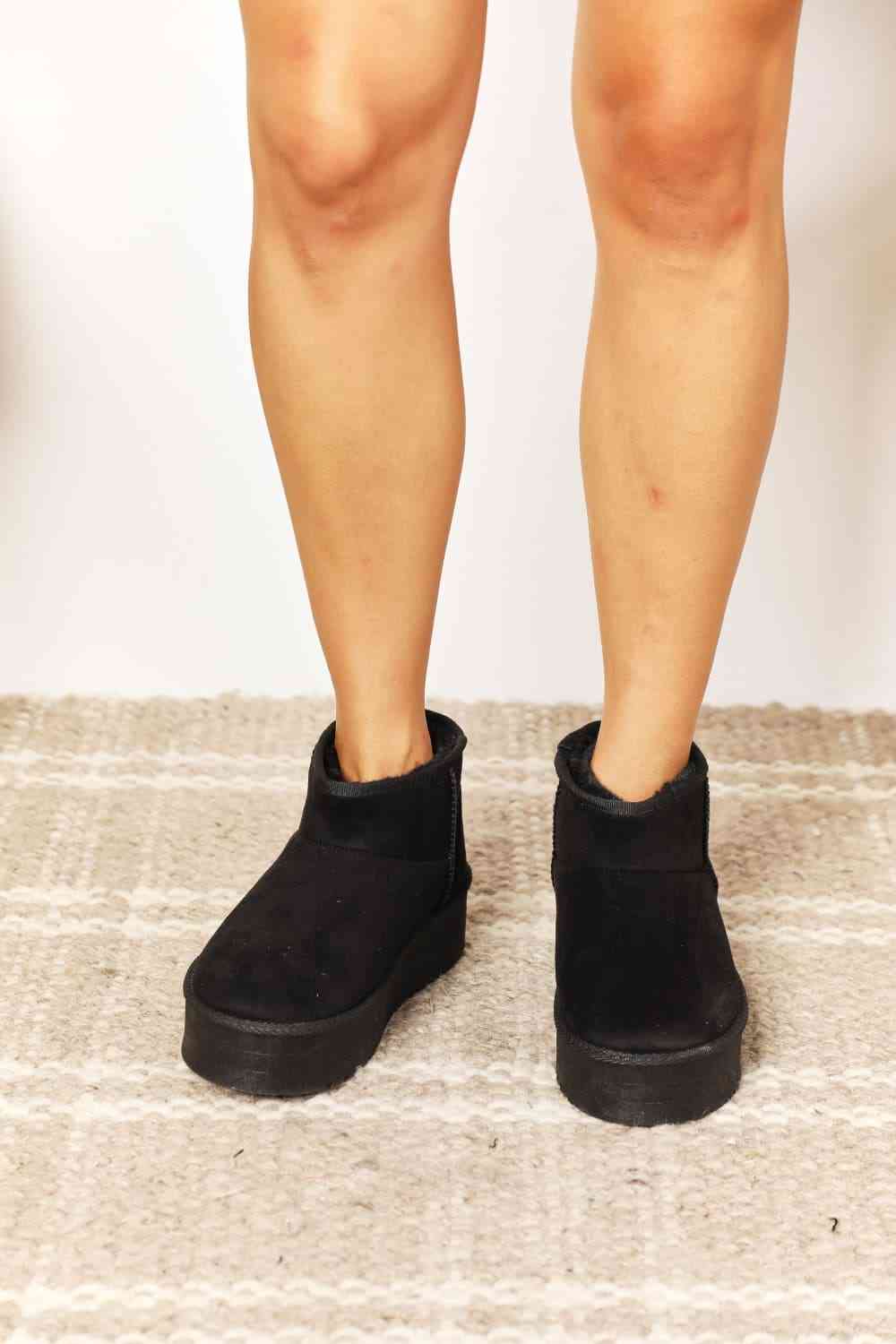 A person is standing on a light-colored patterned rug, wearing Trendsi's Legend Women's Fleece Lined Chunky Platform Mini Boots in black vegan suede.