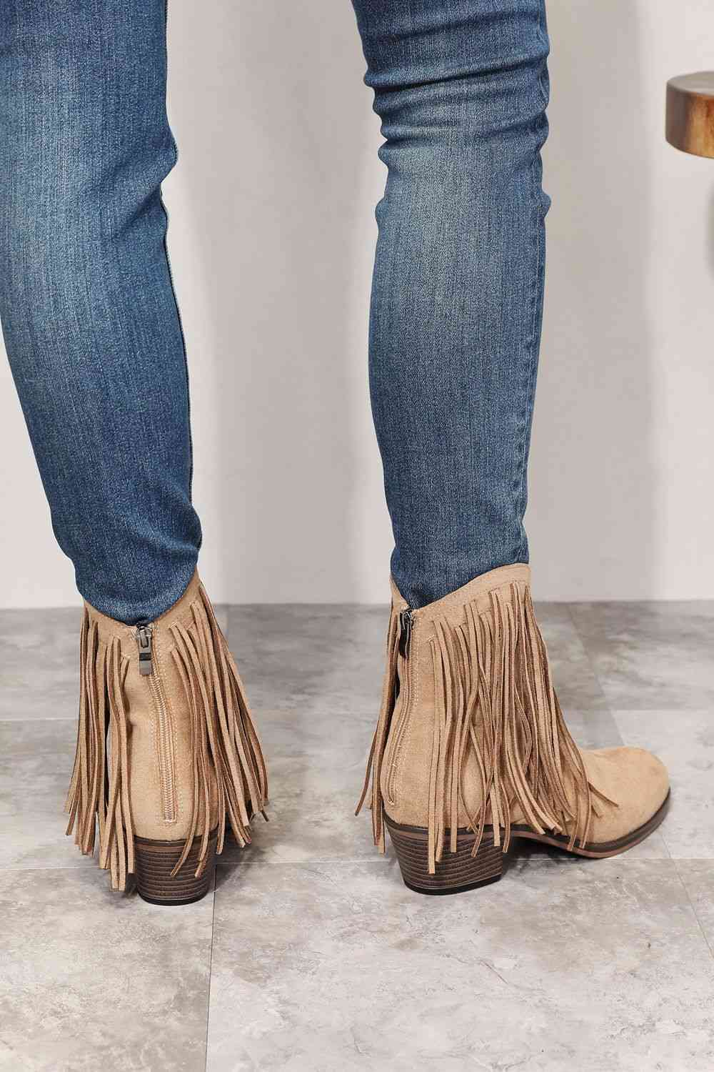 A person stands wearing blue jeans and beige Trendsi Legend Women's Fringe Cowboy Western Ankle Boots on a tiled floor.