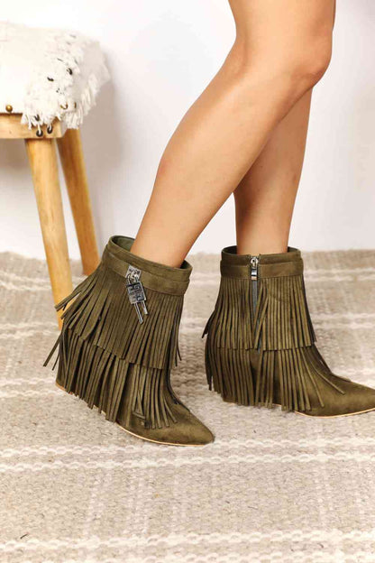A person wearing green Legend Women's Tassel Wedge Heel Ankle Booties with fringe details, standing beside a wooden stool with a white throw.