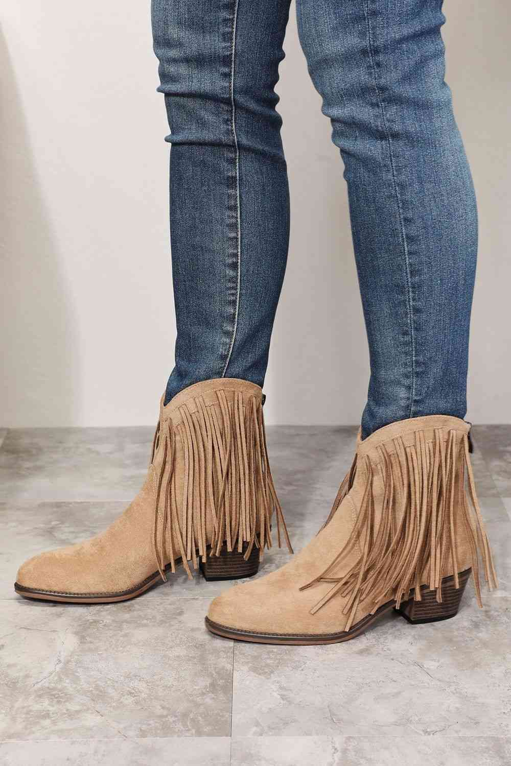 A person standing in beige Trendsi Legend Women's Fringe Cowboy Western Ankle Boots with long fringe details, paired with blue skinny jeans, against a grey tiled floor and white wall background.
